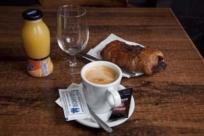 Desayuno sobre una mesa de madera con una taza de café, un pastel, una botella de zumo y un vaso vacío.