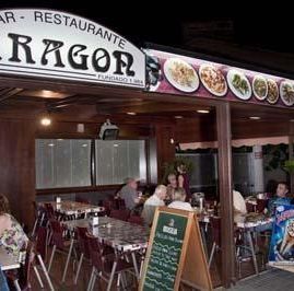 Una terraza de restaurante por la noche, con clientes sentados en mesas bajo un toldo de madera con un letrero que dice 