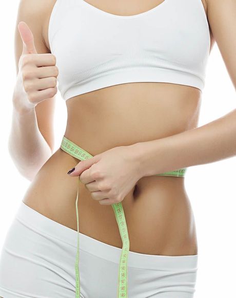 Una mujer vestida con ropa deportiva blanca mide su cintura con una cinta métrica y levanta el pulgar.