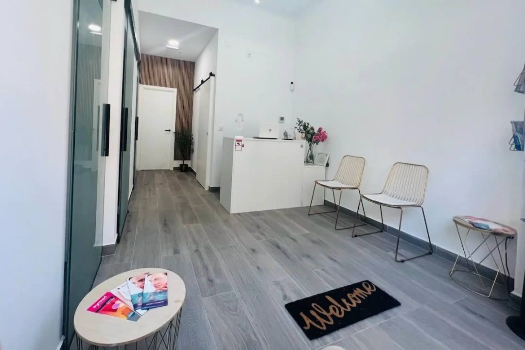 Recepción con escritorio blanco, sillas y suelo imitación madera. Tapete de bienvenida y artículos decorativos.