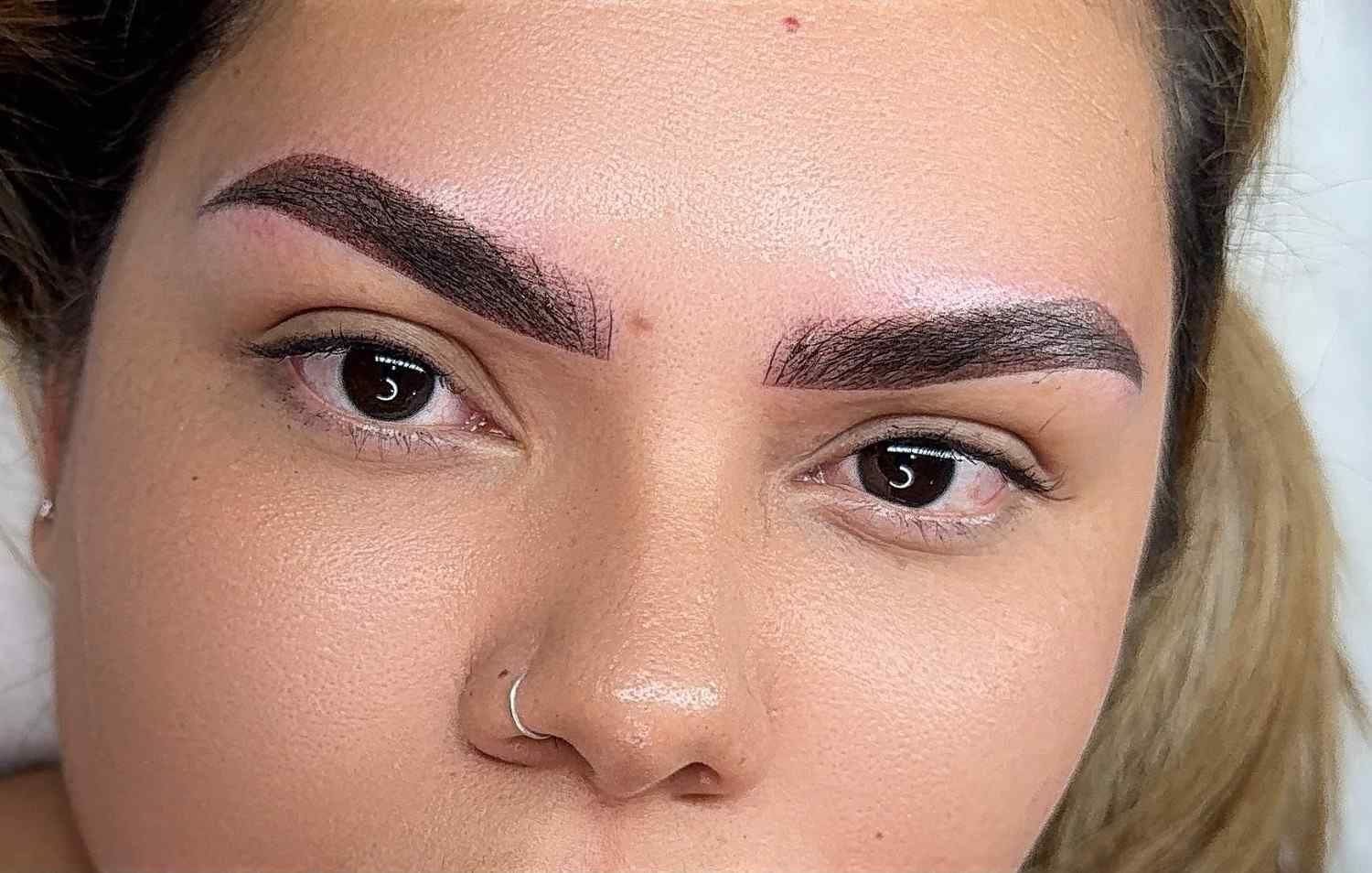 Primer plano de un rostro con cejas oscuras, un piercing en la nariz y maquillaje natural sobre un fondo claro.