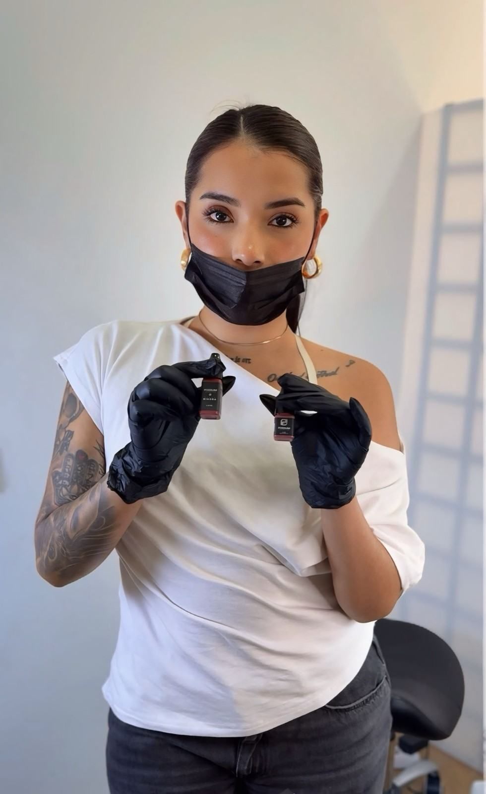 Mujer con guantes negros y mascarilla sosteniendo una máquina de tatuar, de pie en un estudio.