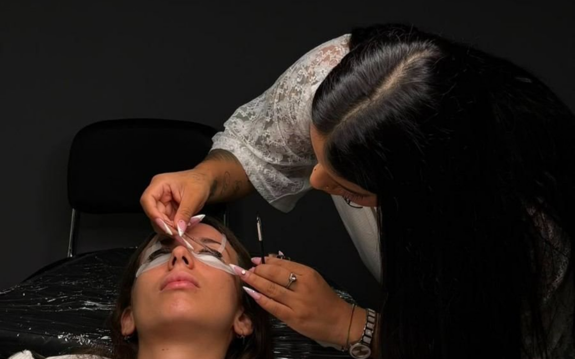 Persona aplicando extensiones de pestañas a una clienta tumbada en una camilla oscura.