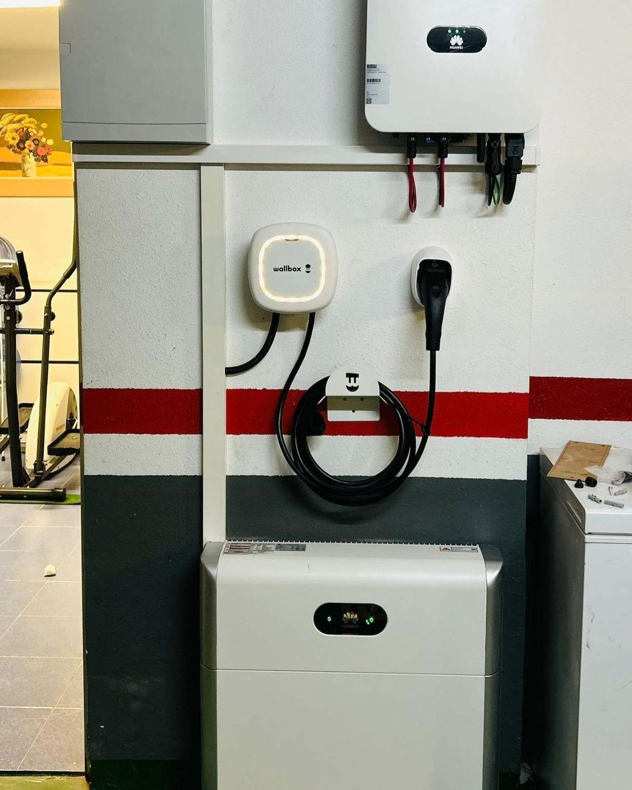 Equipo eléctrico montado en la pared y toma de corriente sobre una pared blanca con una franja roja, con un cable negro enrollado debajo.