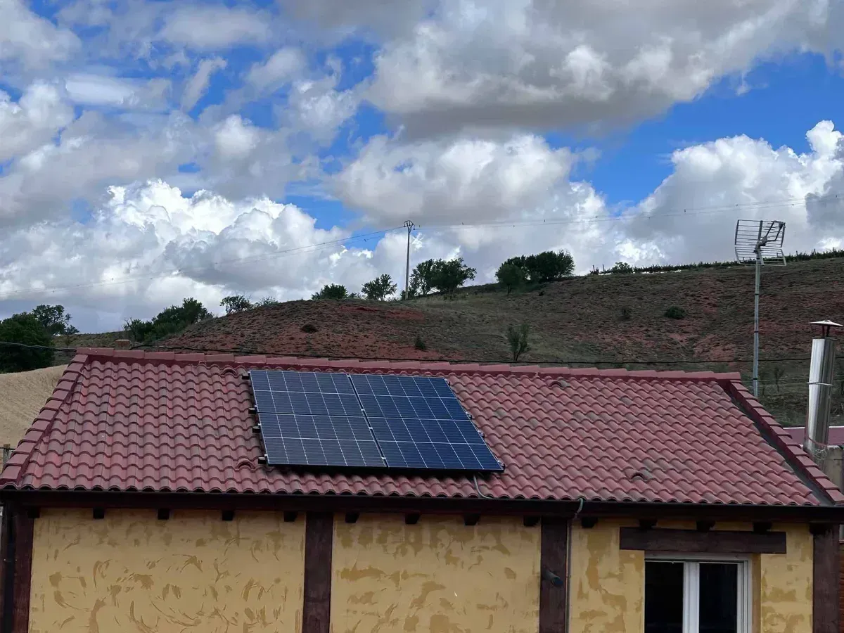 Casa con tejado de tejas rojas y panel solar bajo un cielo nublado, con una colina al fondo