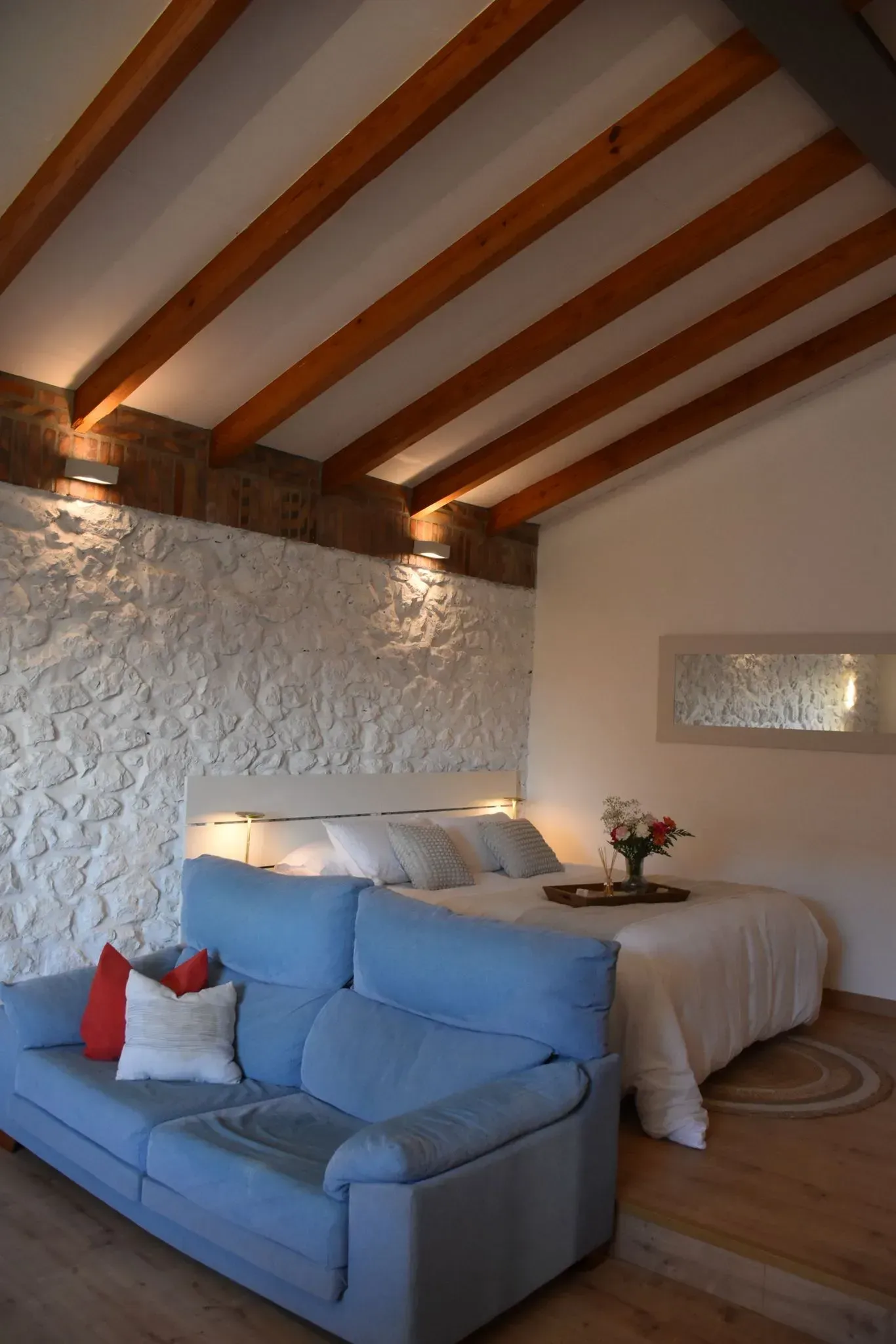Sala de estar con pared de piedra, vigas de madera, mueble rojo y sofá azul.