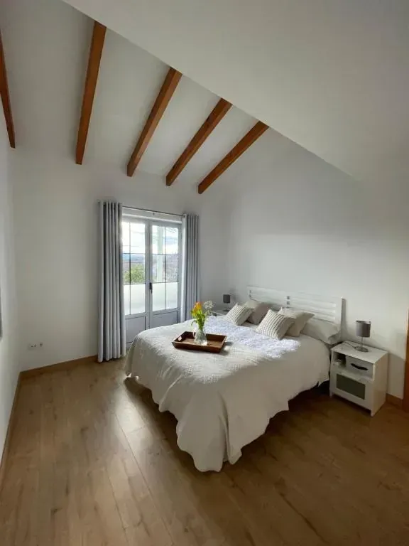Dormitorio con paredes blancas, suelo de madera y vigas vistas. Una cama con sábanas blancas da a una ventana.