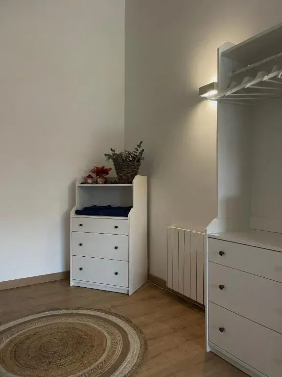 Cómoda blanca con cajones y cambiador en una habitación de tonos claros. Sobre ella hay una cesta de flores.