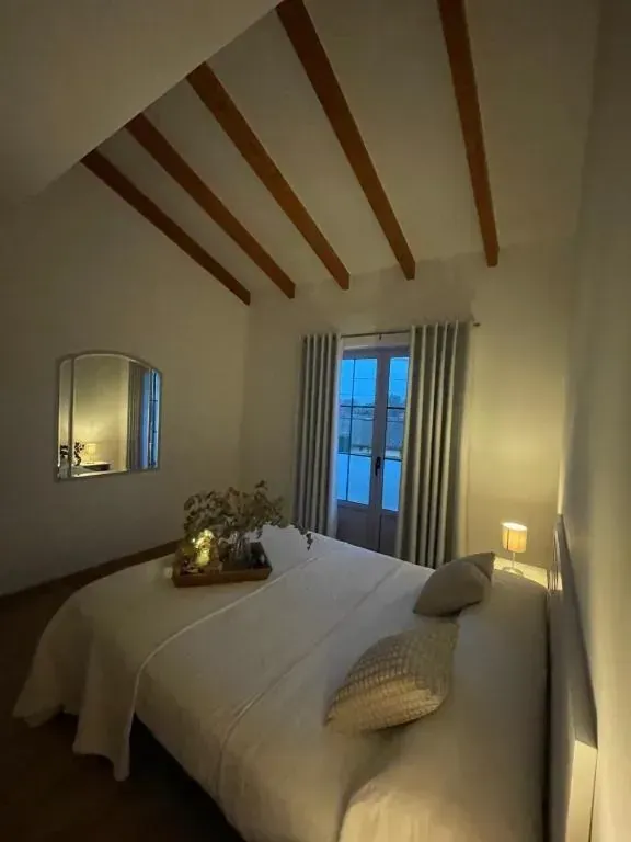 Dormitorio con cama, espejo y ventana con cortinas. Vigas de madera en el techo. Iluminación tenue.