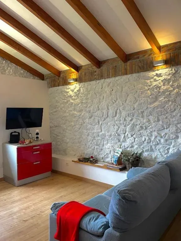 Sala de estar con pared de piedra, vigas de madera, mueble rojo y sofá azul.