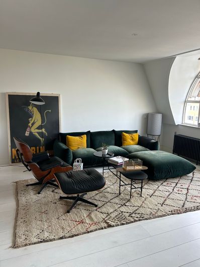 Sala de estar moderna con sofá seccional verde oscuro, dos sillones, alfombra grande y póster vintage.