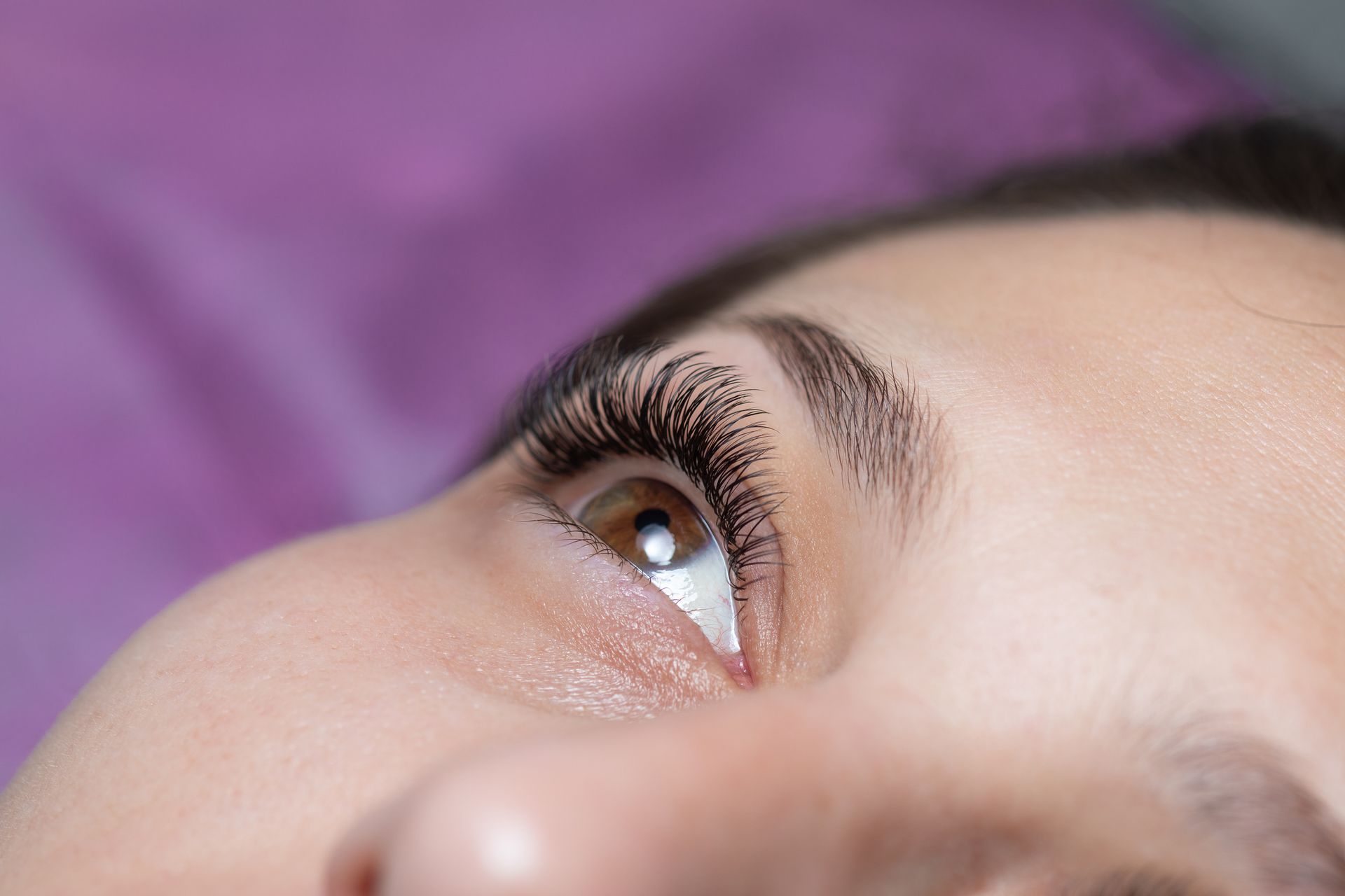 Primer plano del ojo de una mujer con extensiones de pestañas oscuras y densas, mirando hacia arriba. Ojo marrón sobre fondo morado.