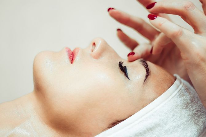 Mujer recibiendo masaje facial; primer plano muestra el rostro con los ojos cerrados, esmalte de uñas rojo y toalla blanca.