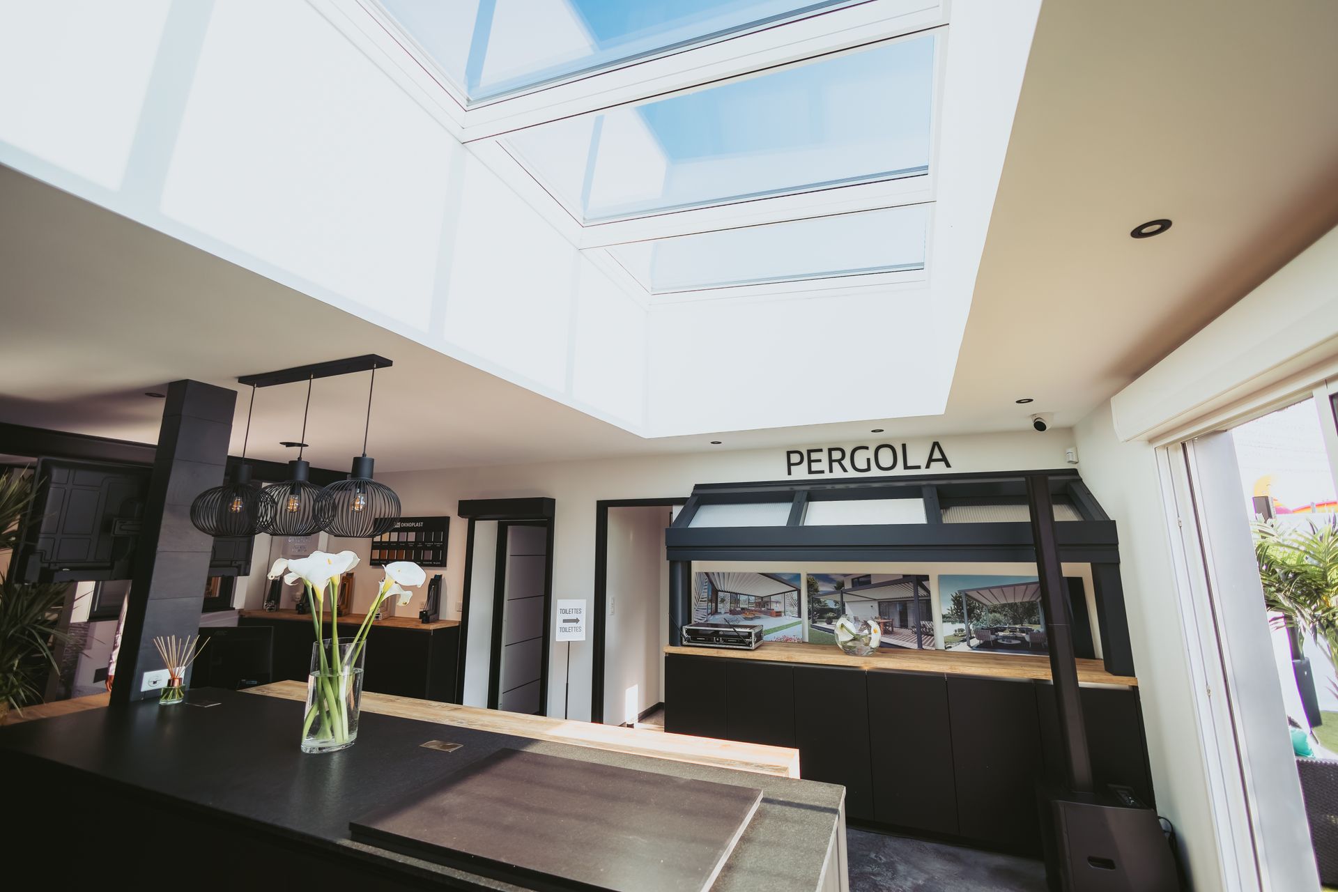 Intérieur de cuisine moderne et ouvert, avec un grand puits de lumière, des armoires foncées, un îlot central et des luminaires suspendus.