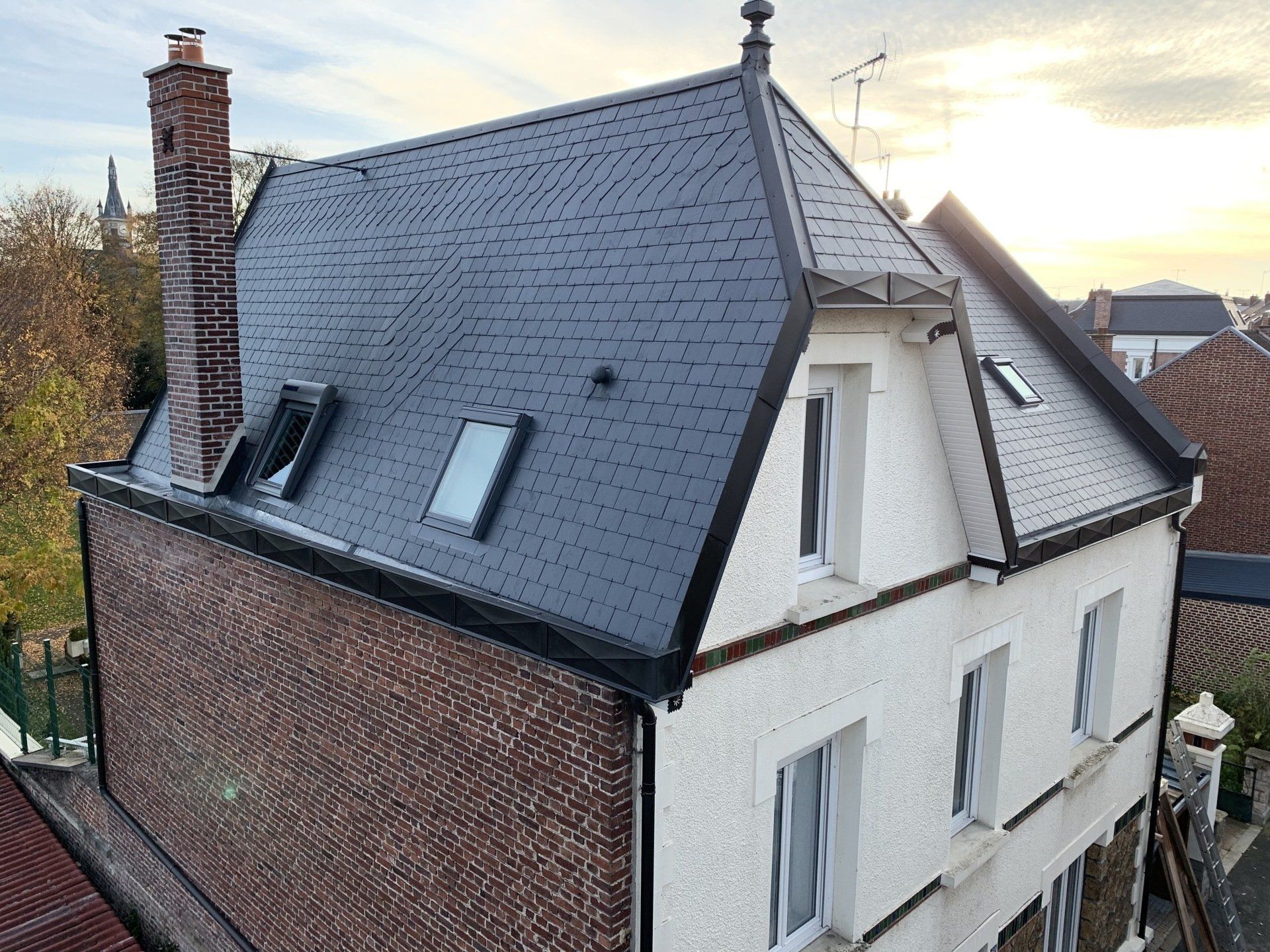 Vue en plongée d'une maison avec un toit en ardoise sombre, une cheminée en briques, des murs en stuc blanc et plusieurs fenêtres de combles.