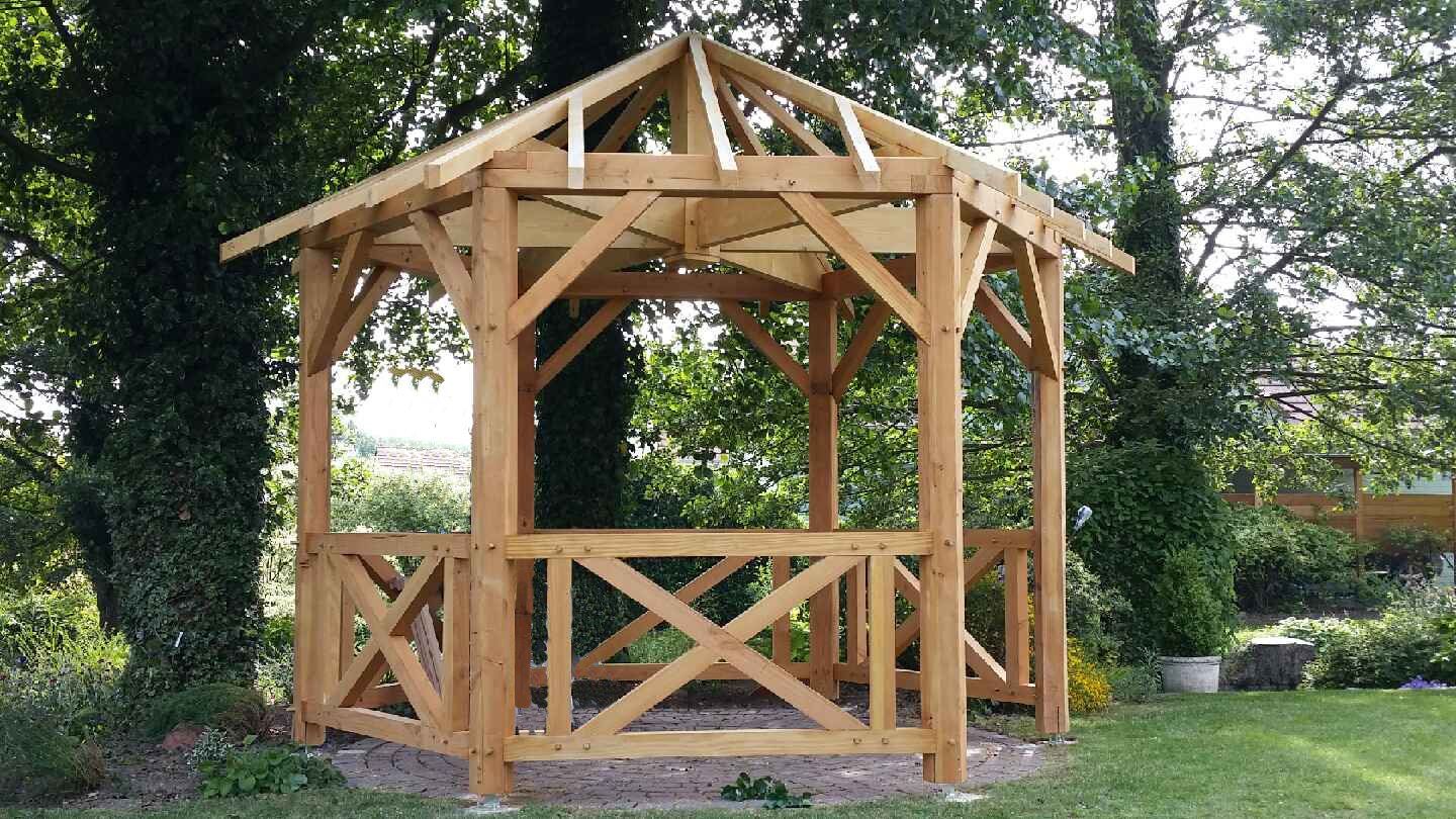 Une structure de gazebo hexagonale en bois avec une rambarde à entretoises, installée dans un jardin verdoyant à l'arrière.