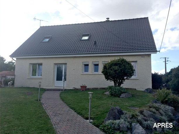 Une maison beige d'un étage et demi avec un toit de tuiles grises, une allée pavée et un jardin paysager sous un ciel nuageux.