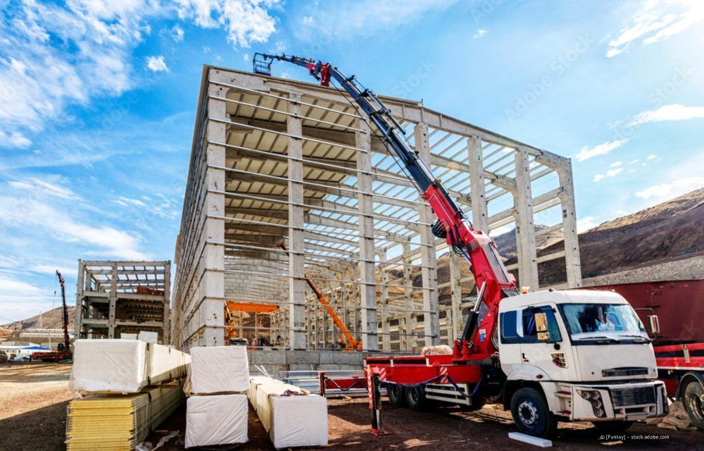 Ein roter, auf einem LKW montierter Kran hebt einen weißen Betonfertigteilträger auf das Dach eines im Bau befindlichen Rohbaus.