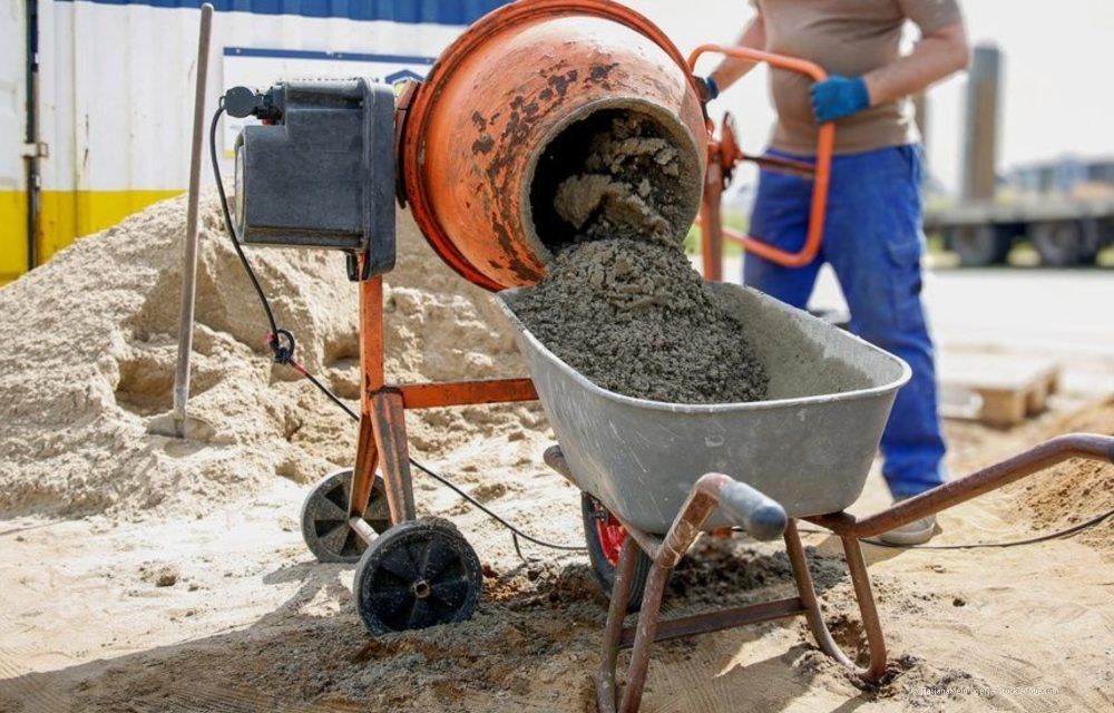 Auf einer Baustelle schüttet ein Arbeiter nassen Zement aus einem orangefarbenen elektrischen Betonmischer in eine graue Schubkarre.