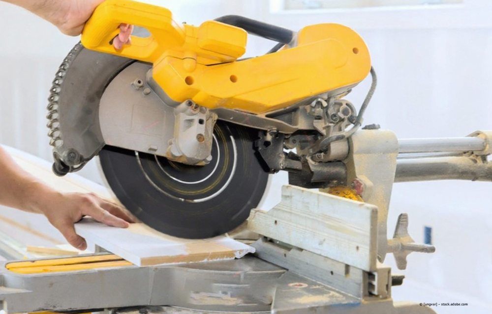 Nahaufnahme von den Händen einer Person, die mit einer gelben Gehrungssäge ein Stück weiße Leiste auf einer Werkbank zuschneidet.