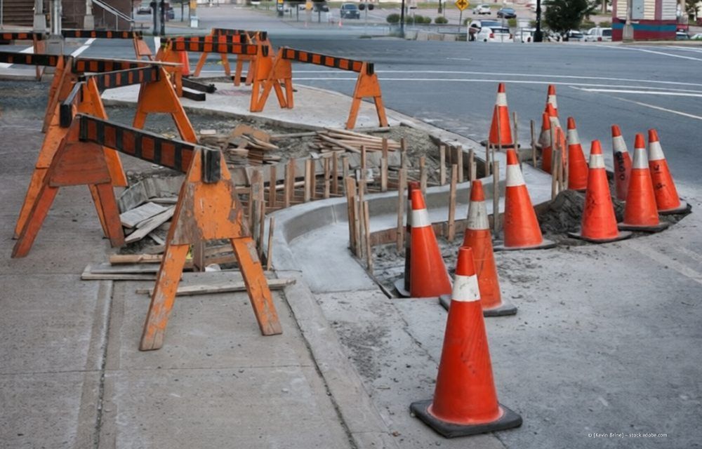 Orangefarbene Sicherheitskegel und Holzbarrikaden sperren einen im Bau befindlichen Abschnitt eines Betonfußwegs und Bordsteins ab.