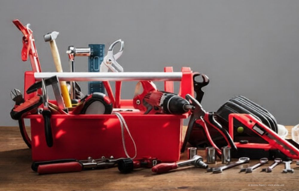 Ein roter Werkzeugkasten aus Kunststoff, gefüllt mit verschiedenen Werkzeugen, darunter Bohrmaschine, Schraubenschlüssel und Hämmer, steht auf einer Holzoberfläche.