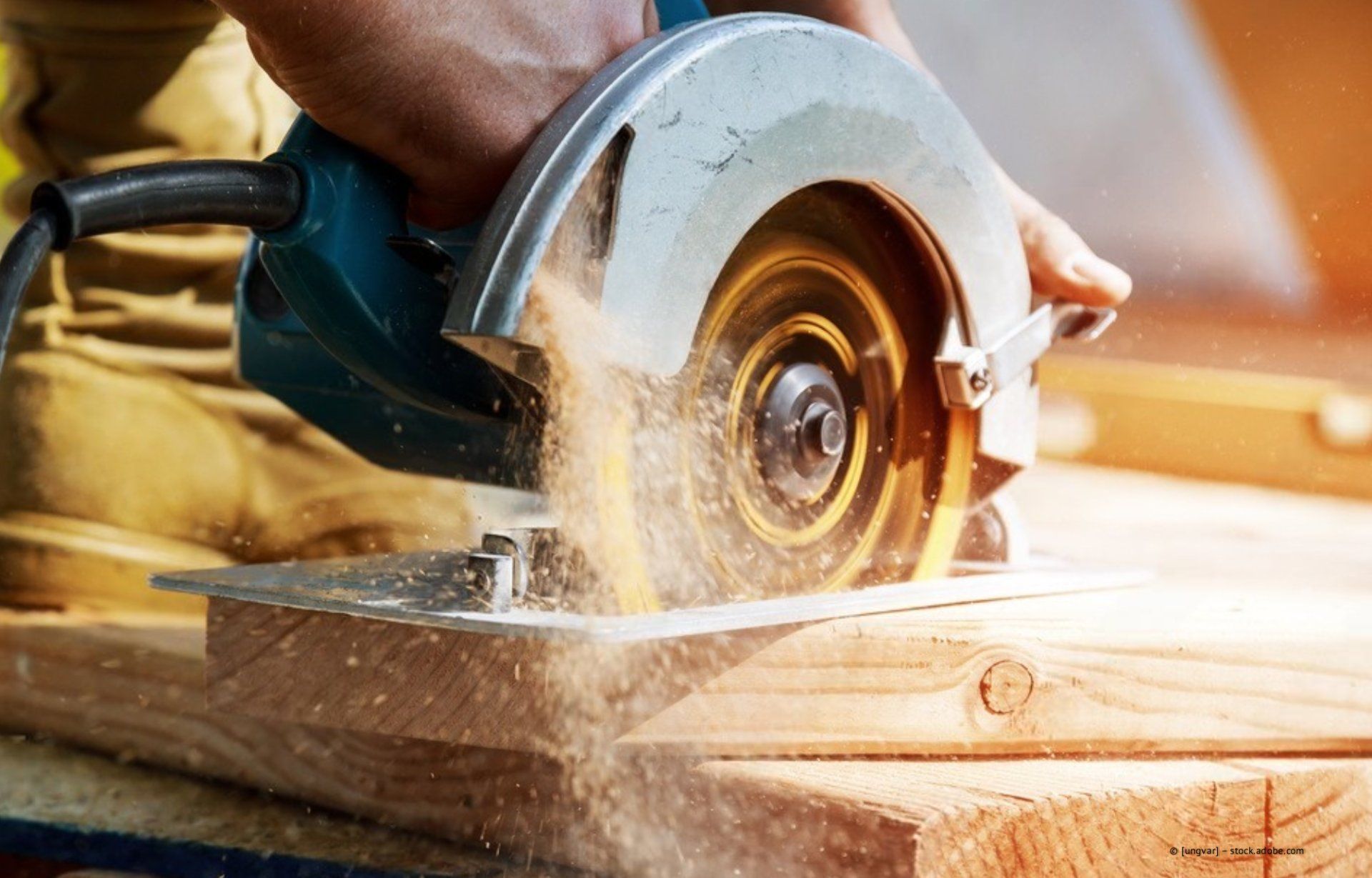 Eine Person schneidet mit einer türkisfarbenen Kreissäge ein Holzbrett, wobei sichtbarer Sägestaub entsteht.