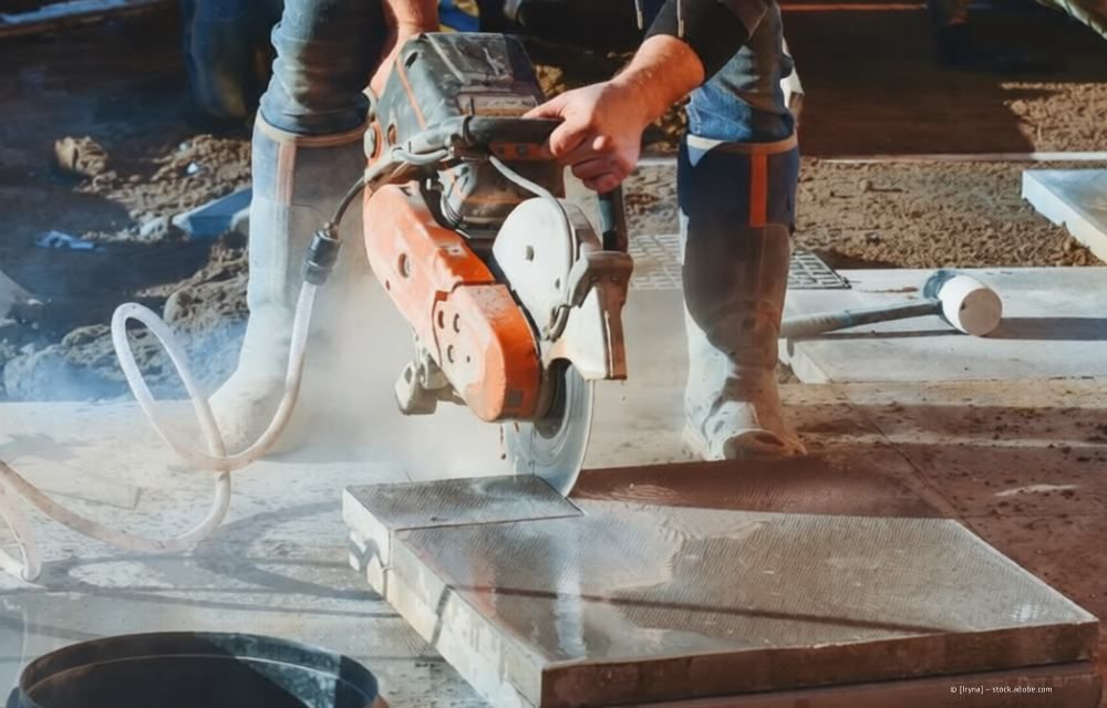 Eine Person benutzt eine handgeführte Betonsäge, um auf einer staubigen Baustelle eine Steinplatte zuzuschneiden.