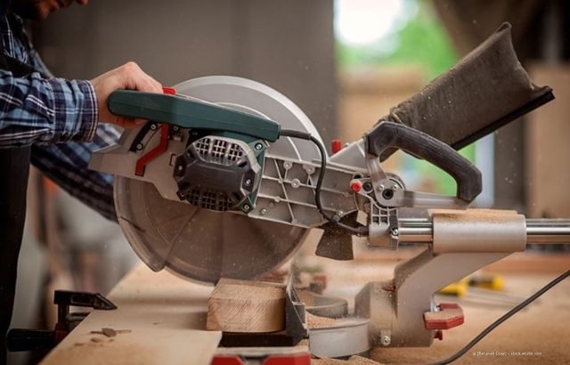 Eine Person in einem karierten Hemd benutzt in einer Werkstatt eine Gehrungssäge, um ein Stück Holz zuzuschneiden.