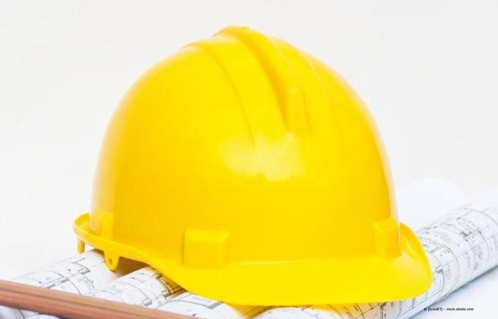 Ein leuchtend gelber Schutzhelm liegt auf zusammengerollten Architekturplänen.