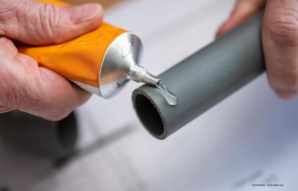 Mit den Händen wird aus einer gelben Tube ein durchsichtiger Klebstoff auf den Rand eines grauen Kunststoffrohrs aufgetragen.