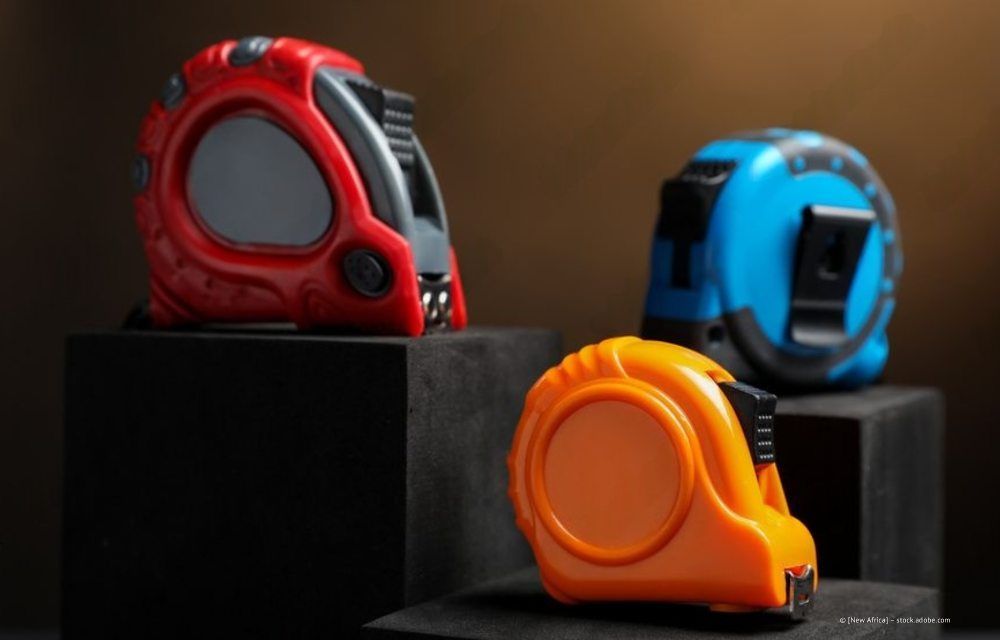 Drei Maßbänder in Rot, Orange und Blau sind auf schwarzen Sockeln vor einem dunklen, düsteren Hintergrund ausgestellt.