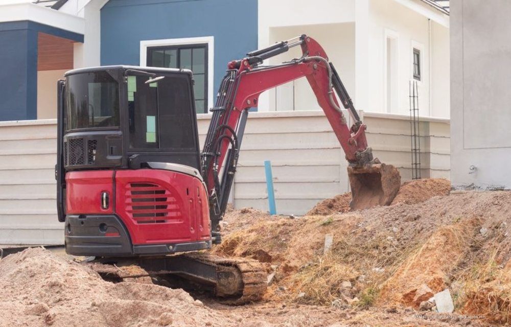 Ein kleiner roter Bagger steht auf einem Erdhaufen neben einem im Bau befindlichen Haus.