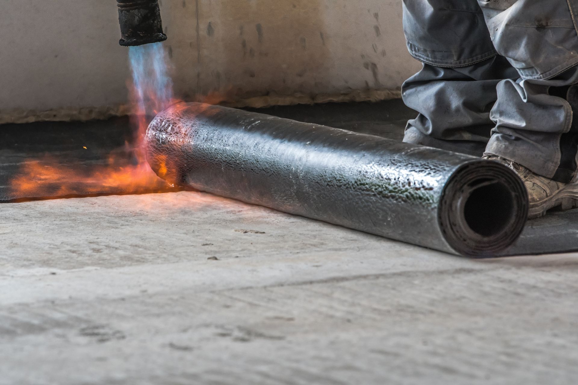 Un ouvrier soude un rouleau noir sur un sol en béton ; des étincelles et une flamme sont visibles.