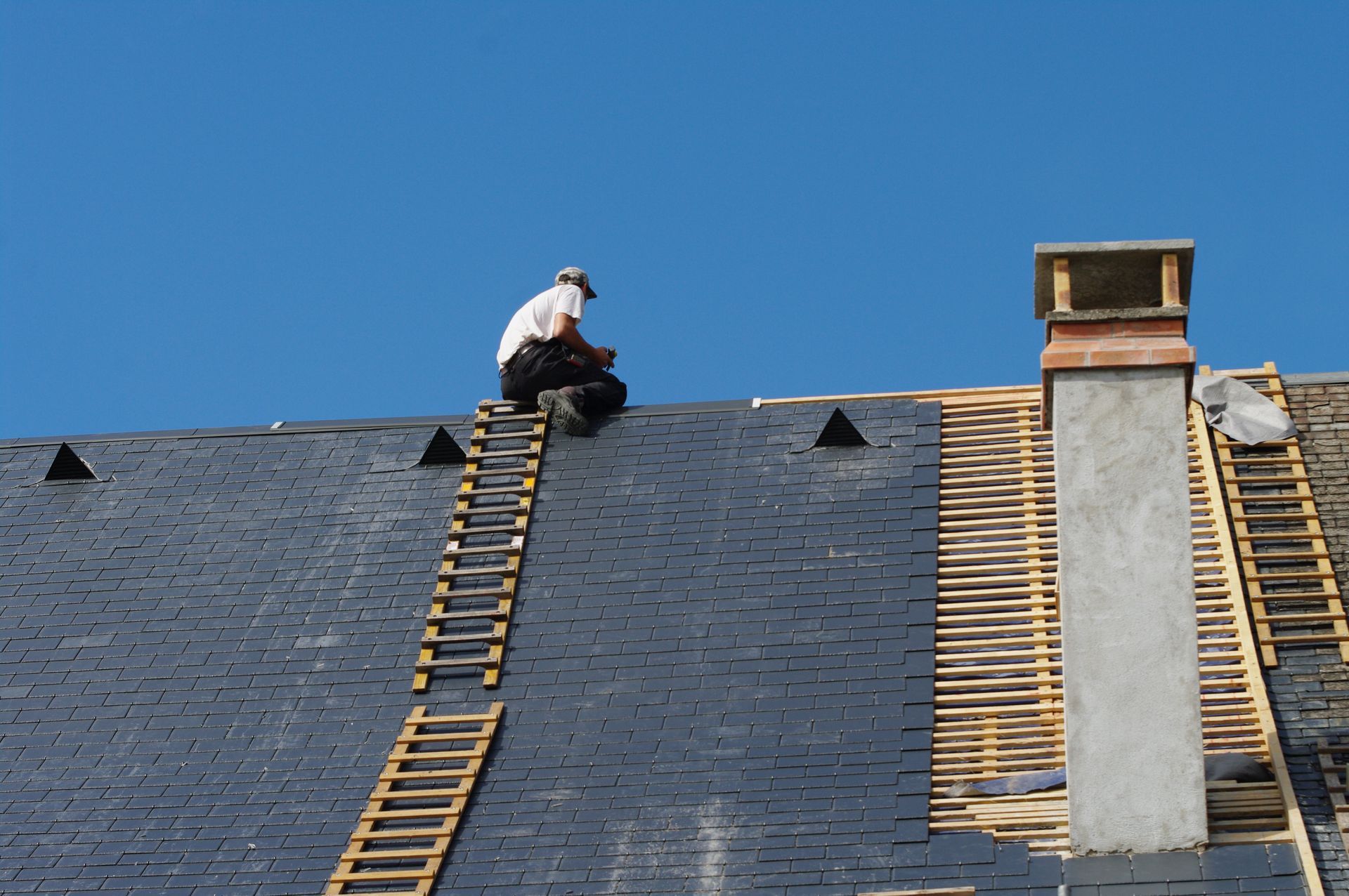 Ouvrier sur un toit, près d'une échelle et d'une cheminée, sous un ciel bleu clair.