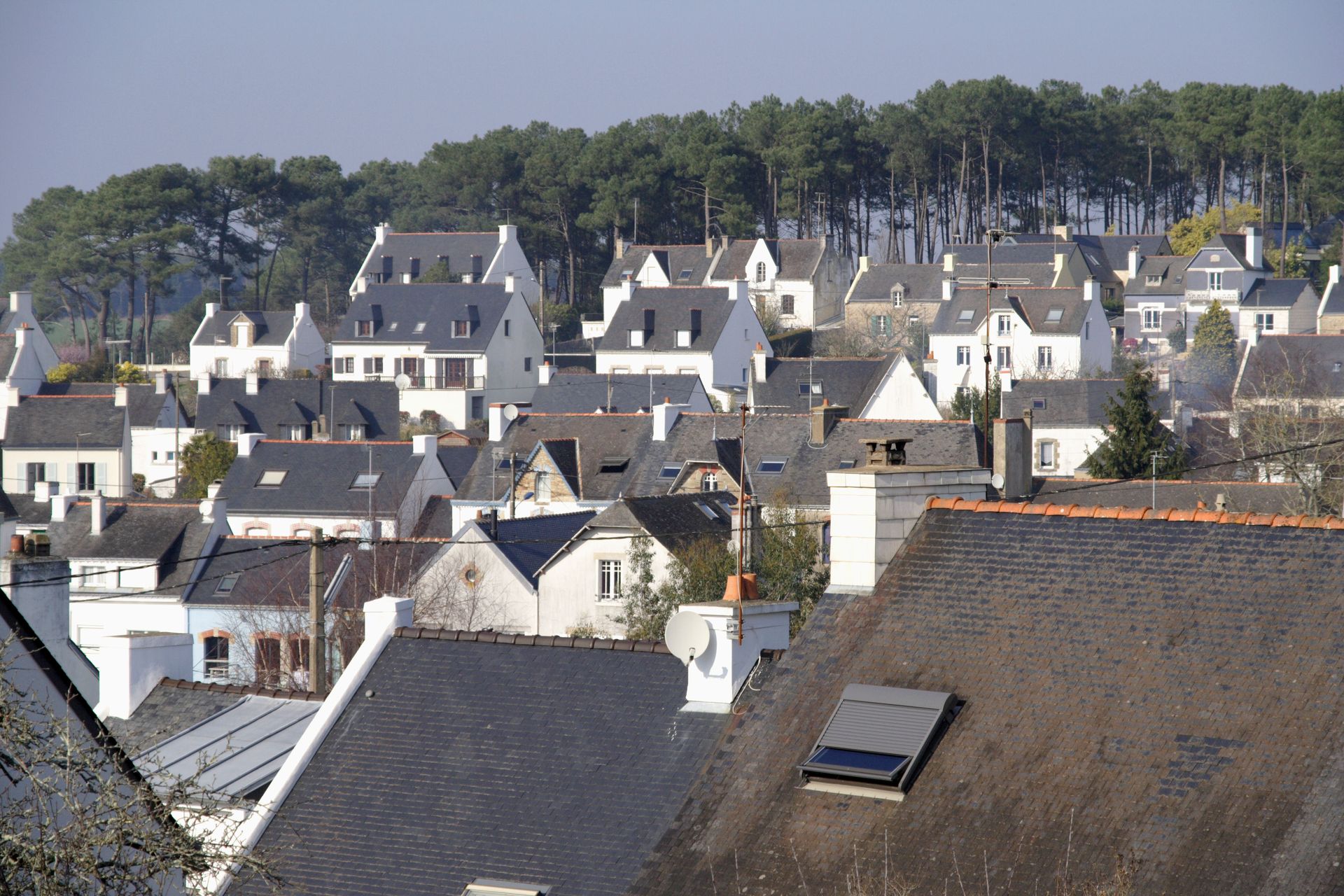Un village à flanc de colline, avec des rangées de maisons blanches aux toits gris, sous une crête boisée.
