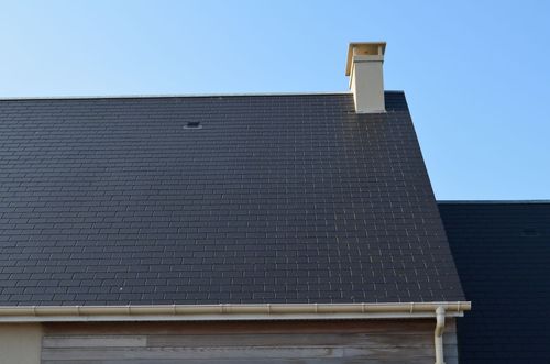 Toit en ardoises sombres avec une cheminée blanche se détachant sur un ciel bleu clair
