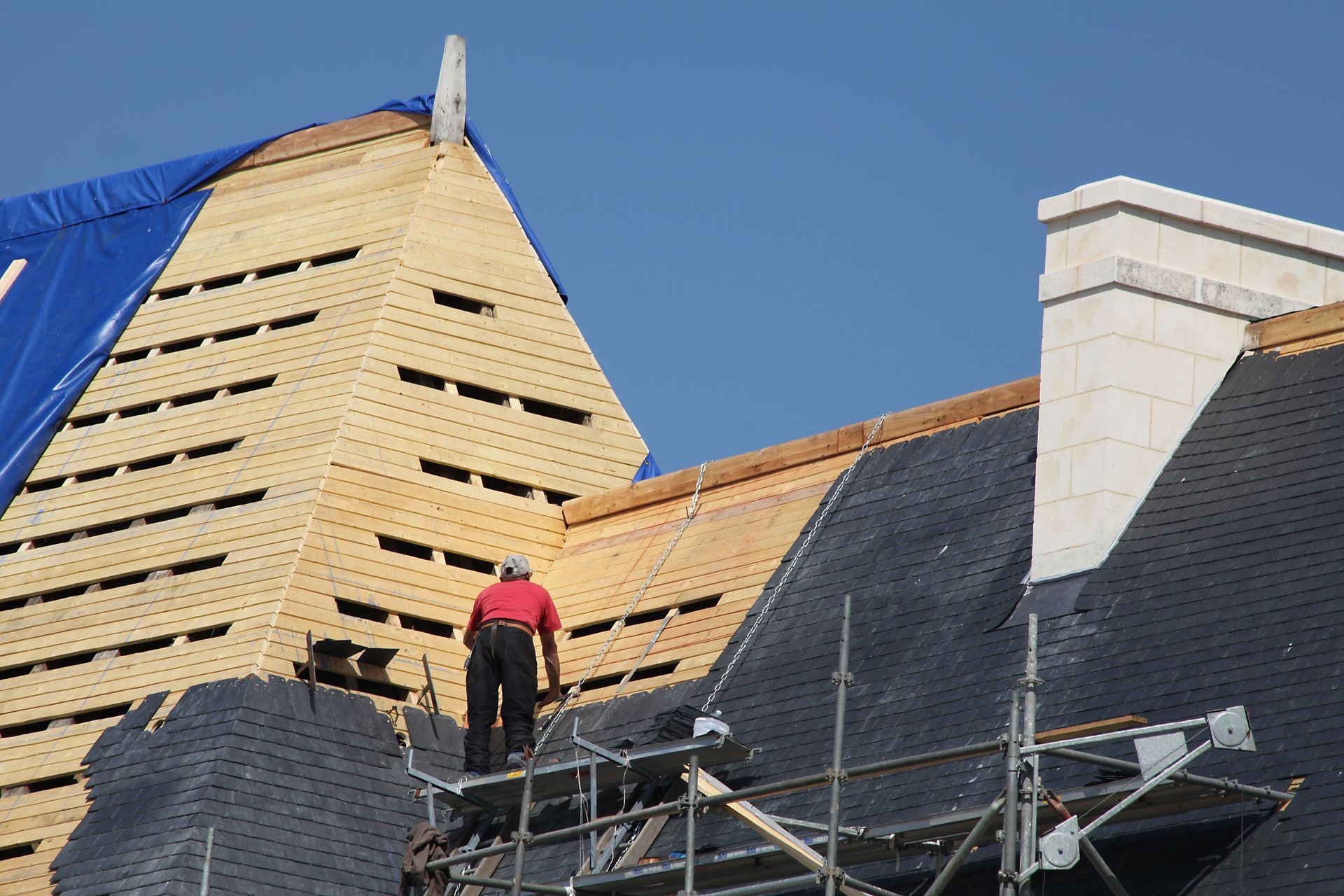 Des couvreurs travaillent sur un toit pentu en construction, avec une bâche bleue et des échafaudages se détachant sur un ciel dégagé.