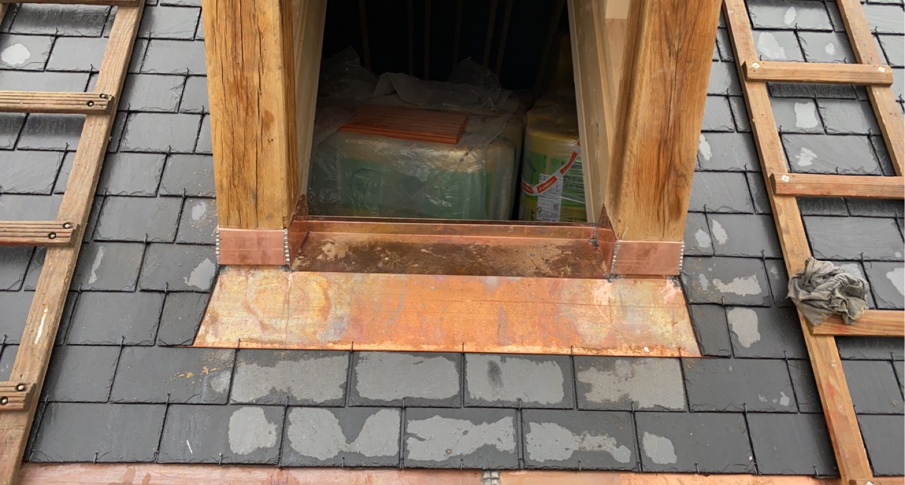 Fenêtre de toit à cadre en bois ouvert, entourée d'ardoises grises et d'une base en cuivre.