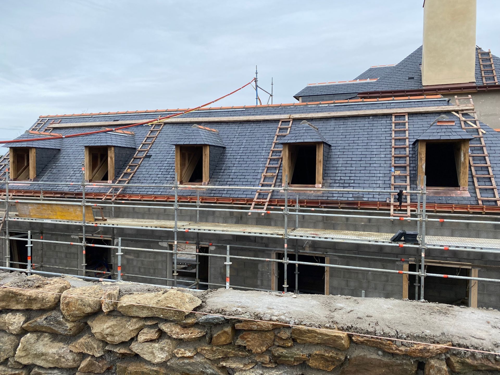 Maison en construction avec toit d'ardoise, échafaudages et mur de pierre devant.