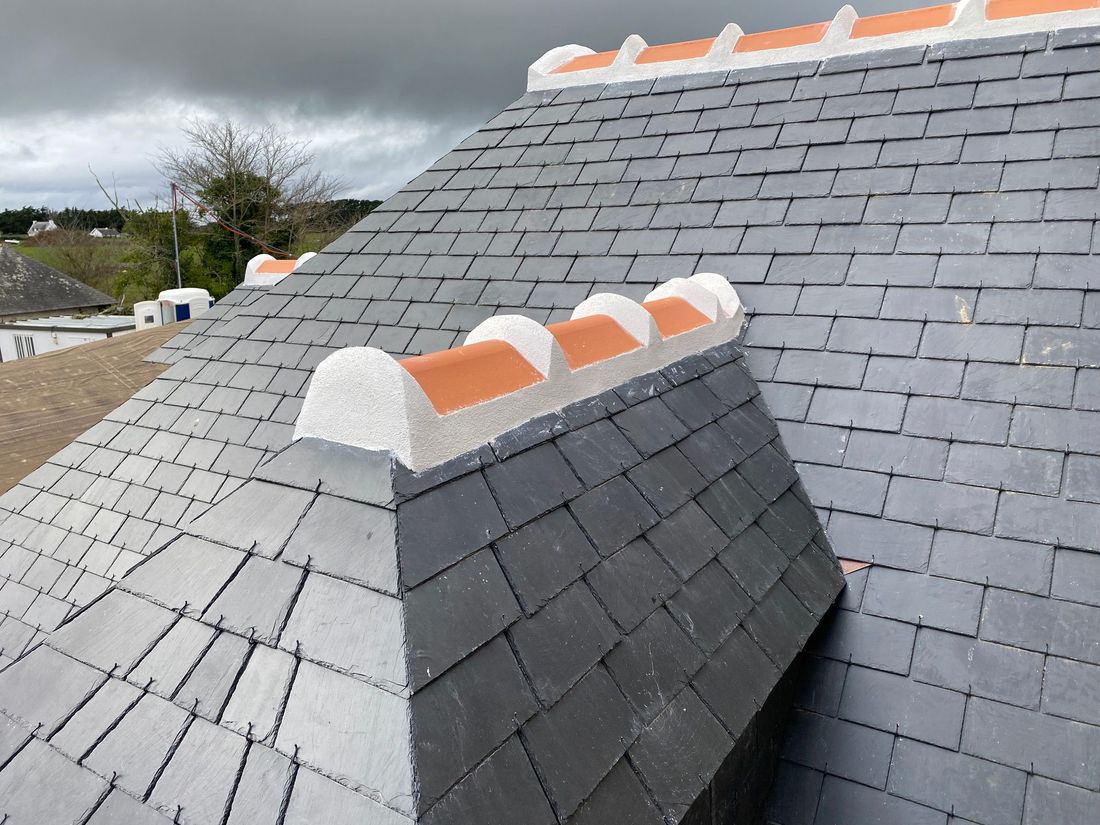 Toit en ardoise grise avec des tuiles faîtières orange sous un ciel nuageux