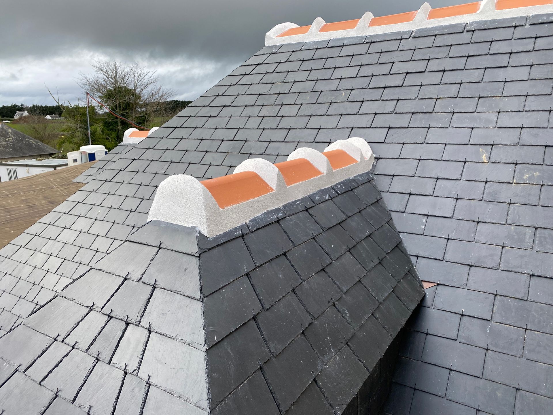 Toit en ardoise avec des tuiles faîtières blanches et orange sous un ciel nuageux