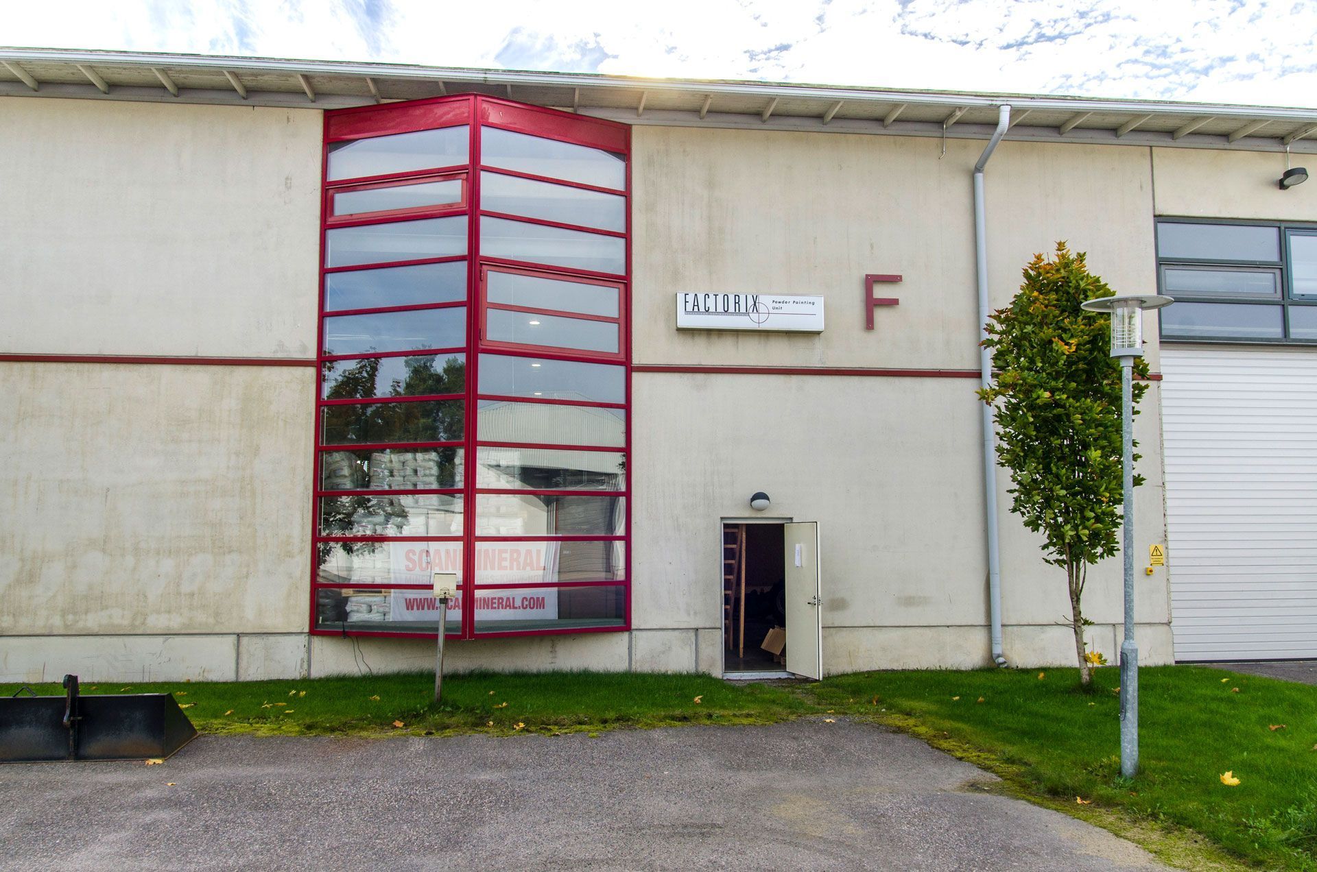 Rakennuksen ulkoasussa punakehyksiset ikkunat ja "F"-kirjaimella merkitty ovi. Vihreää ruohoa ja pieni puu näkyvät.