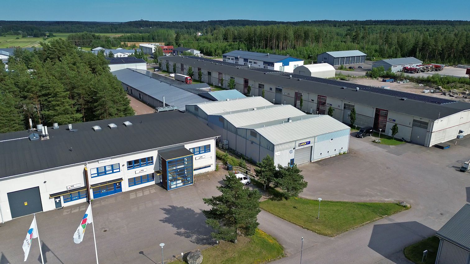 Ilmakuva teollisuuskompleksista, jossa on pitkiä, matalia rakennuksia puiden ympäröimänä, kirkkaan taivaan alla.