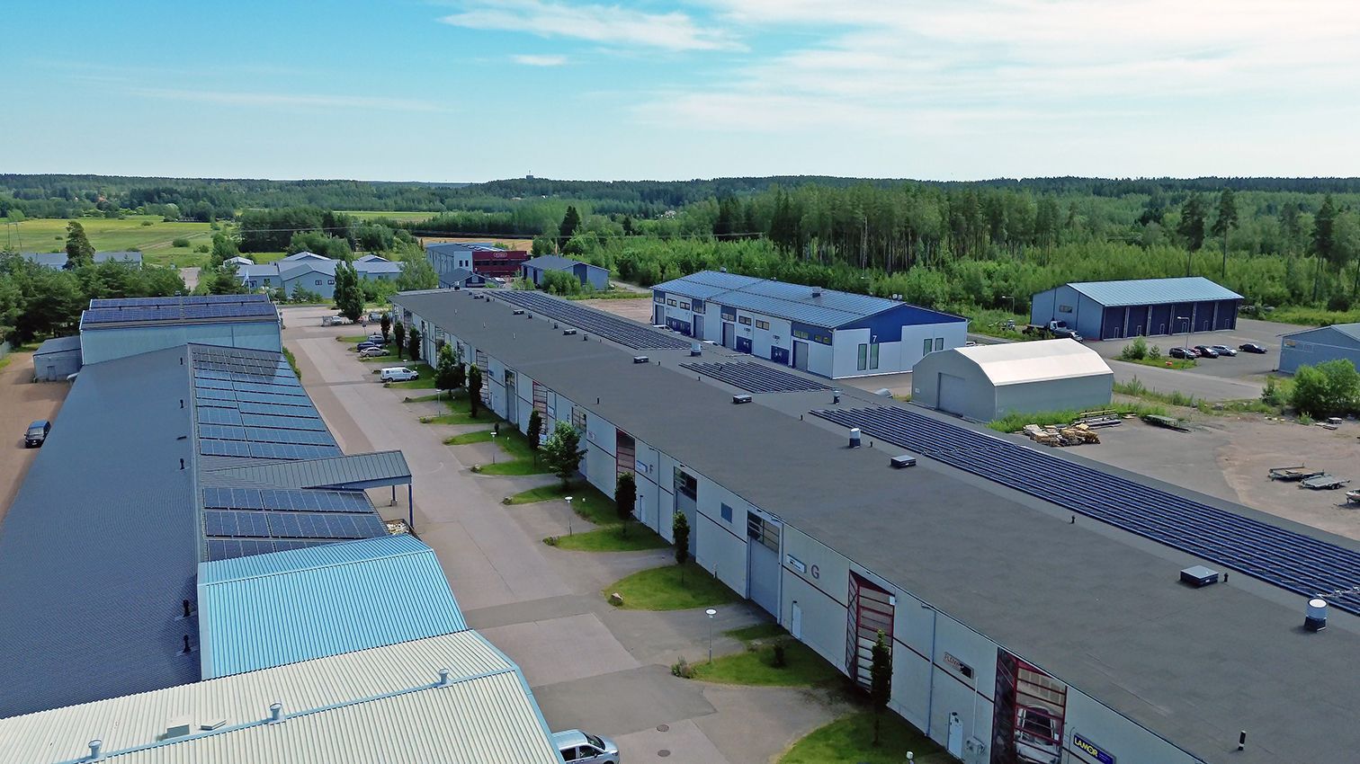 Ilmakuva teollisuusrakennuksista, joissa on pitkät, tummat katot. Vihreitä puita ja sininen taivas taustalla.