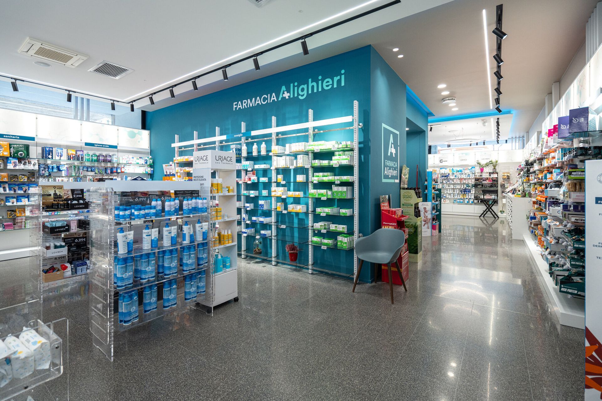 Luminoso corridoio della farmacia con scaffali color verde acqua, espositori di medicinali e alcuni clienti all'interno di un negozio moderno.