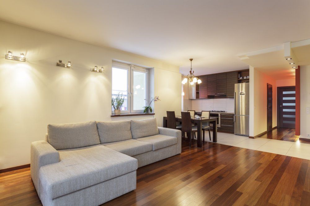 Modern open-plan living room with gray sectional sofa, wooden floors, and kitchen/dining area beyond