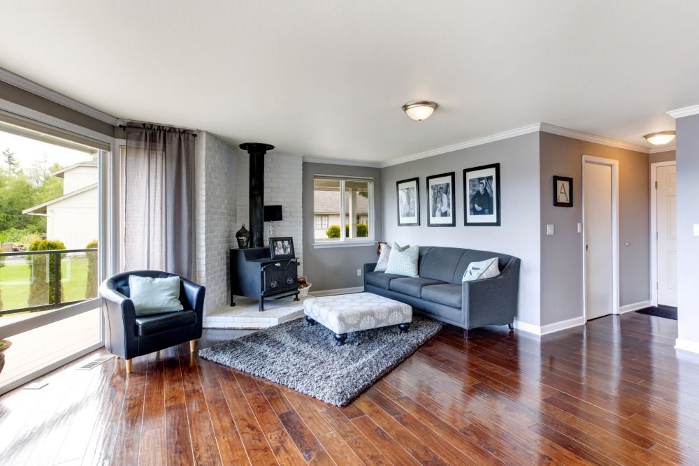 Bright living room with gray sofa, black chair, wood stove, large window, and hardwood floors