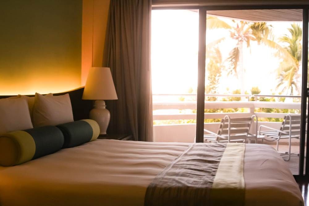 Hotel room with a bed and lamp, overlooking a bright balcony with palm trees