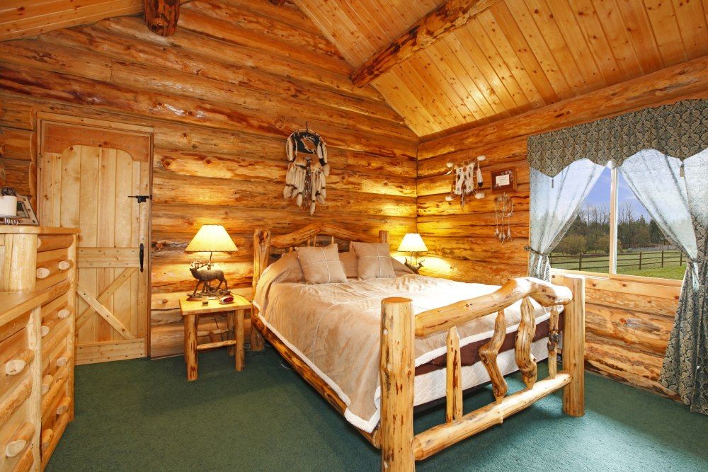 Cozy log cabin bedroom with a wooden bed, warm lamps, and a large window overlooking trees and water